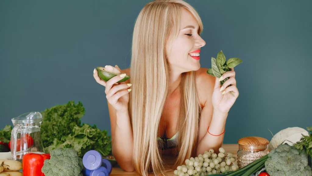 A smiling woman holding avocado and basil, surrounded by fresh vegetables and fruits for healthy eating.